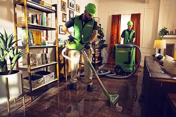 Two SERVPRO technicians in green vests and hard hats extracting standing water from a flooded residential living room using professional water extraction equipment and hoses.
