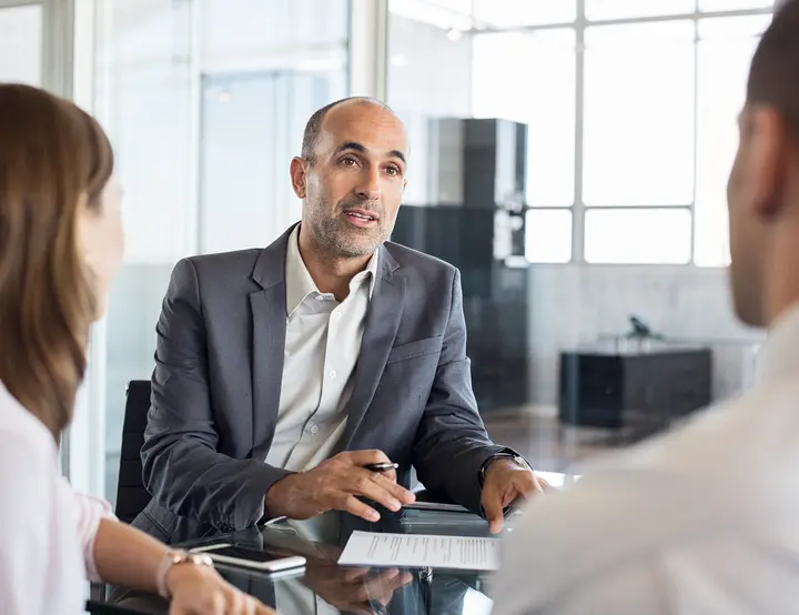 A professional insurance agent or consultant in a gray blazer meeting with two clients across a glass desk in a modern office, reviewing documents and discussing coverage options.